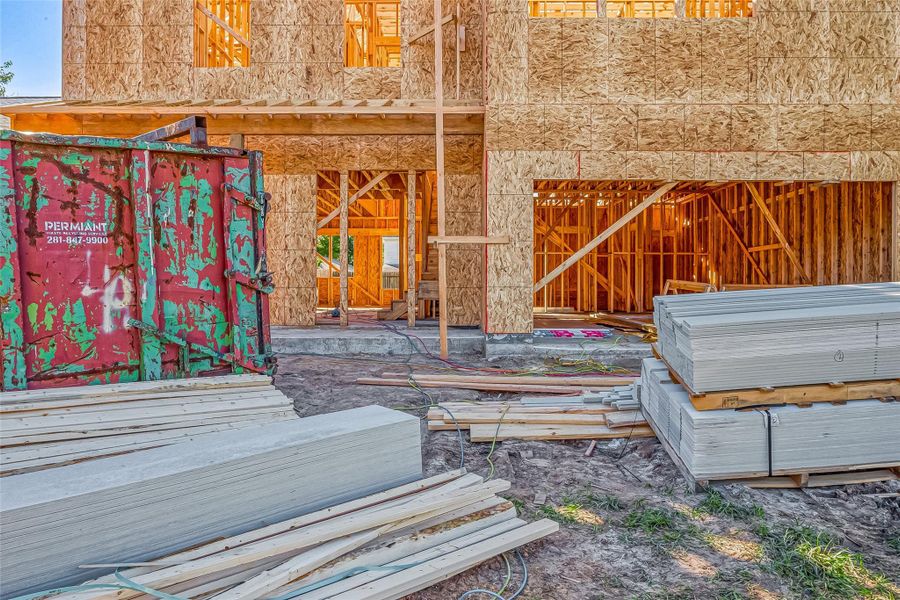 This photo shows a house under construction. The structure is framed with wood and oriented strand board (OSB). Building materials are stacked nearby, and there's a large dumpster on site, indicating ongoing construction work.