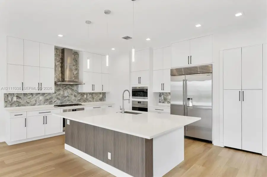 Kitchen featuring crisp white cabinetry, a center island with a ribbed wood-finish base, quartz countertops, a patterned stone tile backsplash, and stainless steel appliances