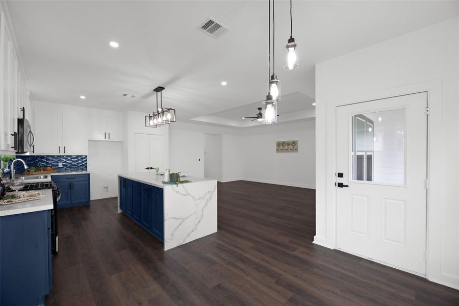 Modern, open-concept kitchen and family area with dark wood floors. The kitchen features blue cabinetry, a sleek white marble island, and stainless steel appliances. Stylish pendant lights and a ceiling fan enhance the bright, airy space.