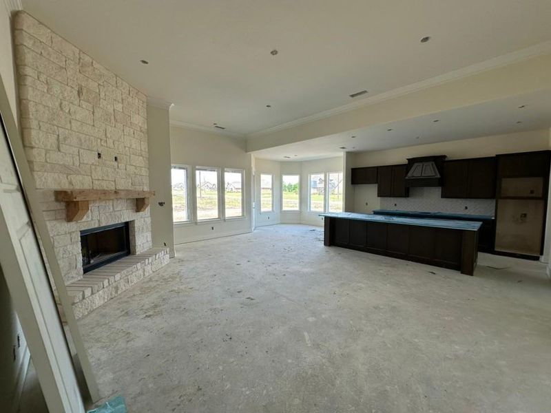 Open-concept living area featuring a stone fireplace with a wood mantle, multiple windows, dark wood cabinetry, a large kitchen island, and crown molding