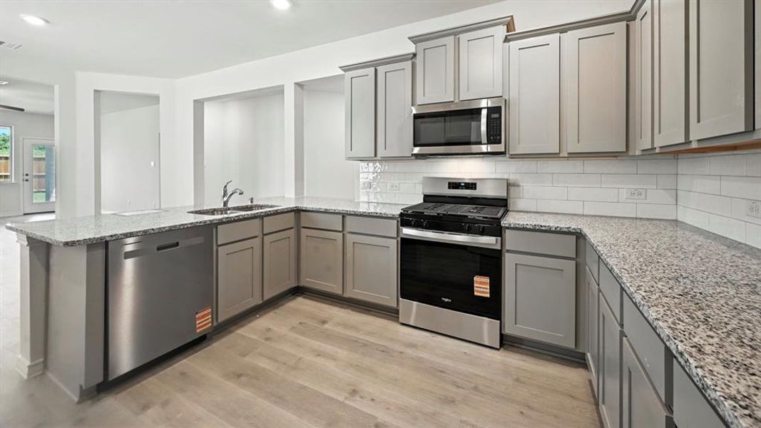 Kitchen with gray cabinetry, stainless steel appliances, decorative backsplash, light stone countertops, and light wood finished floors
