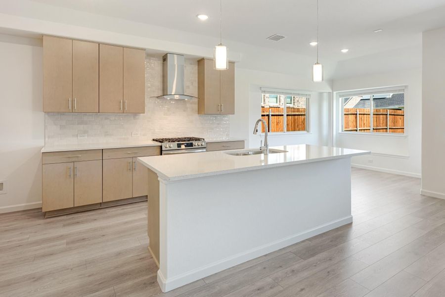 Open kitchen with large center island, light wood cabinets, stainless range, and pendant lights over light gray floors Open kitchen with large center island, light wood cabinets, stainless range, and pendant lights over light gray floors