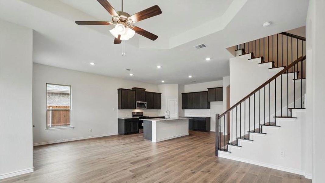 Open-concept interior featuring wood-finish flooring, a contemporary kitchen with dark cabinetry and a white island, a staircase with dark wood handrails and black metal balusters, recessed lighting, and a ceiling fan