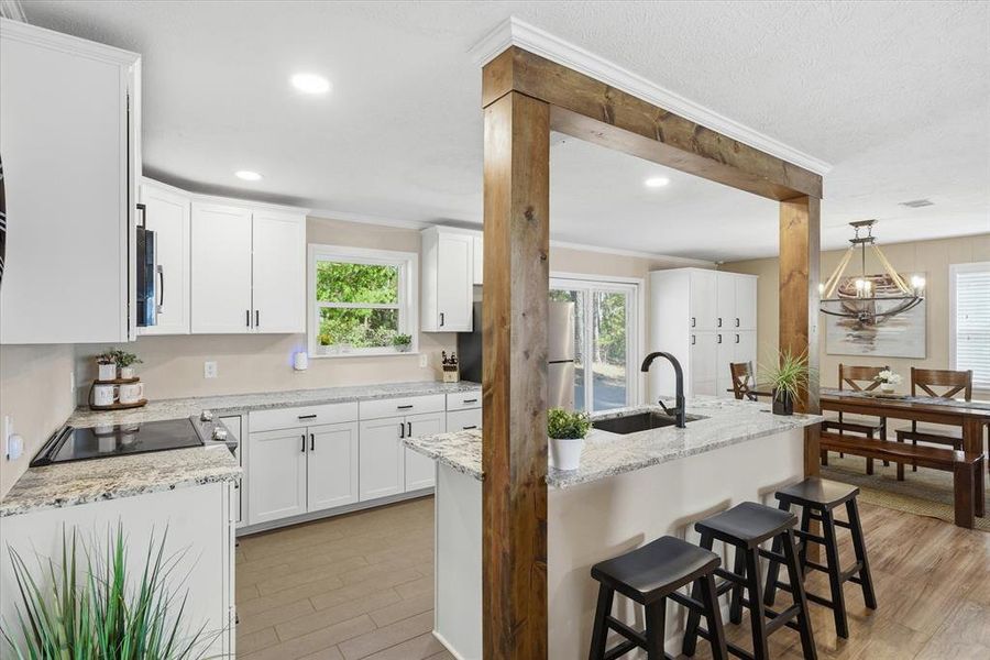 Open-concept kitchen featuring white cabinetry, light-toned granite-style countertops, an integrated countertop stove, a matte black gooseneck faucet, and a large central island with a built-in sink