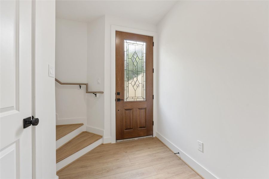 The foyer blends warmth and modern simplicity with a decorative glass front door, light flooring, white walls, and black accents. A nearby staircase leads to the main living level, while the entry area provides a polished, inviting transition into this thoughtfully designed new construction home.