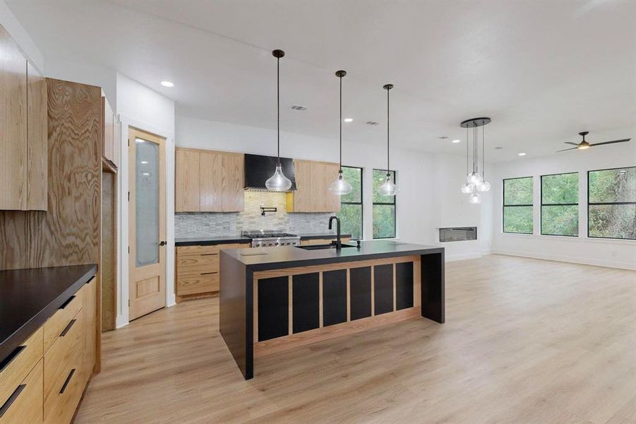 Expansive kitchen and living area featuring a central island with pendant lighting, light wood-finish flooring, a modern fireplace, and multiple large windows Expansive kitchen and living area featuring a central island with pendant lighting, light wood-finish flooring, a modern fireplace, and multiple large windows