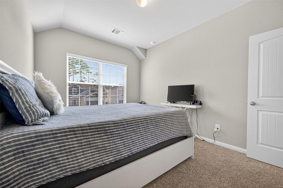 This is a cozy bedroom featuring a large bed with striped bedding, soft carpeting, neutral walls, and a window that lets in natural light. It includes a small desk with a TV, providing a comfortable and functional space.