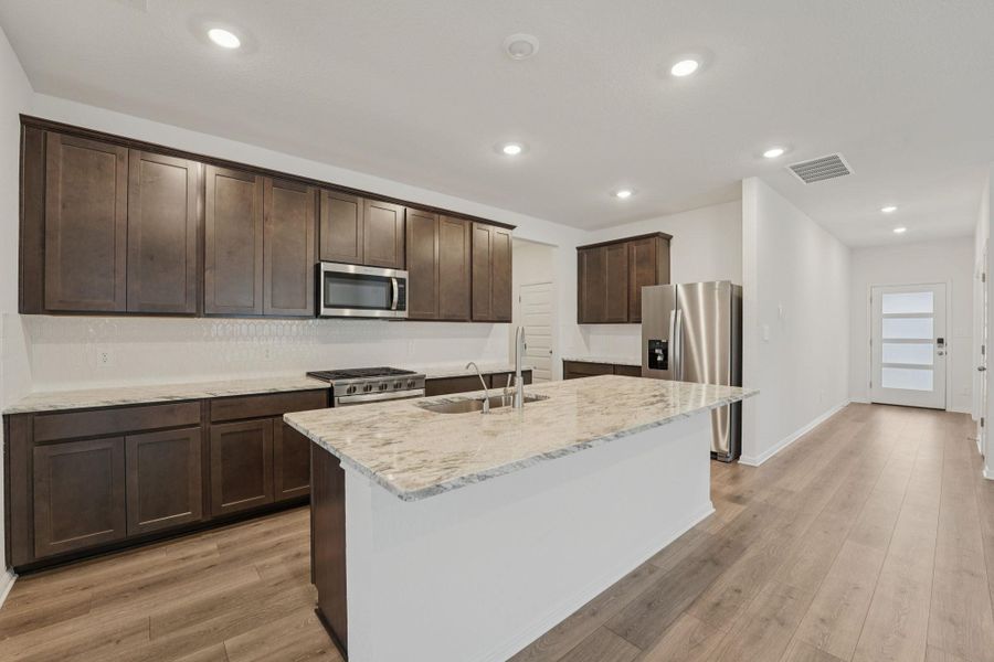 Kitchen with dark brown cabinetry, stainless steel appliances, recessed lighting, light stone countertops, and an island with sink