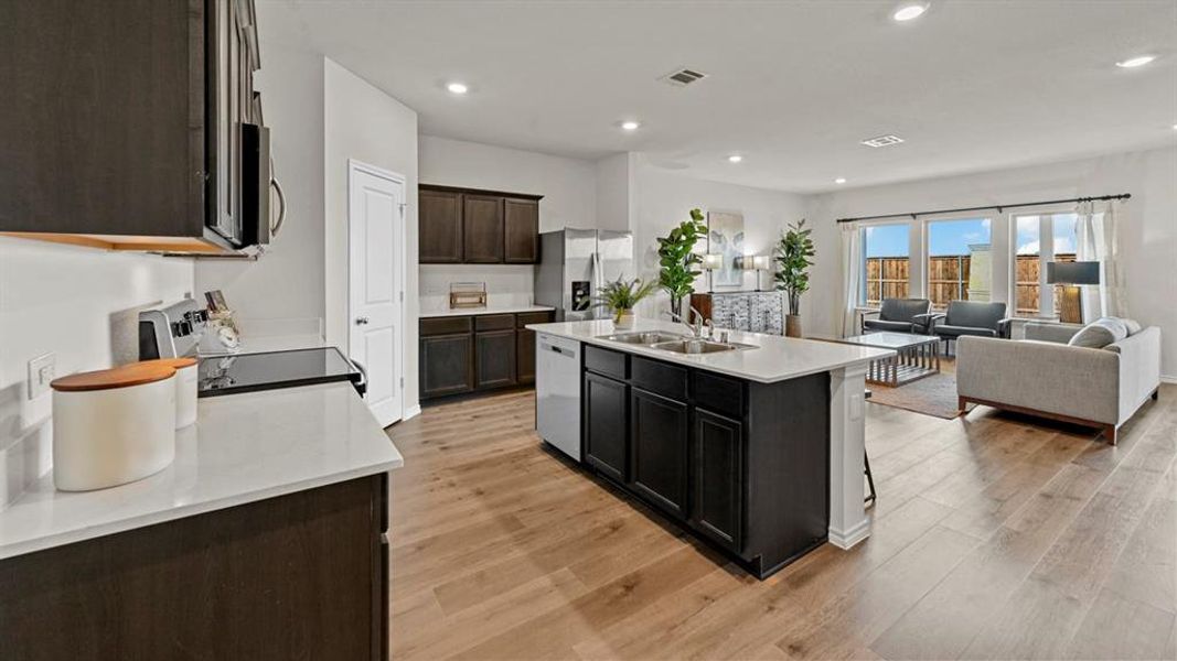 Open concept living area featuring wood flooring, a kitchen island with a sink, stainless steel appliances, and large windows with privacy fencing visible Open concept living area featuring wood flooring, a kitchen island with a sink, stainless steel appliances, and large windows with privacy fencing visible