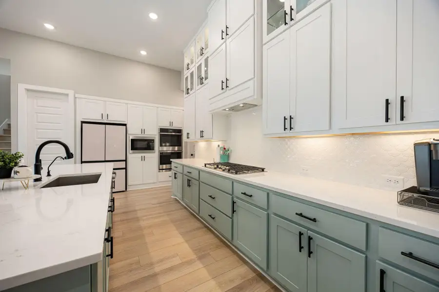 This bright and inviting kitchen showcases a stylish two-tone cabinet design with sleek black hardware and abundant storage, including extended upper cabinets with glass-front detailing. The expansive quartz countertops and large island provide plenty of workspace, complemented by a modern tile backsplash and stainless steel appliances. Warm wood flooring ties the space together, creating a perfect balance of function and elegance.