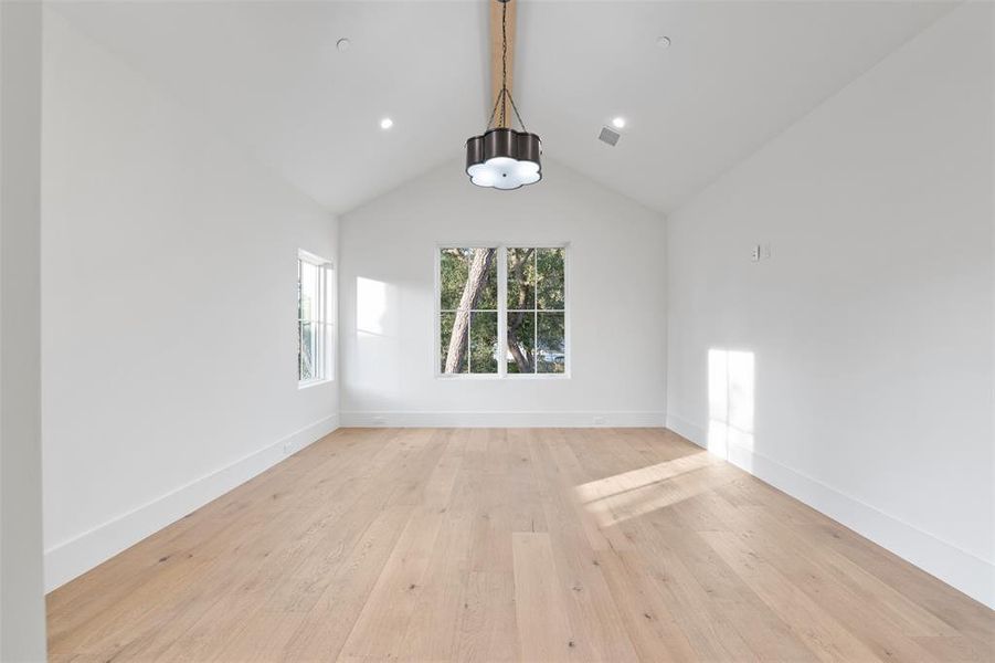 The photo shows a bright, empty room with a vaulted ceiling, modern lighting fixtures, hardwood floors, and a window with a view of greenery outside.