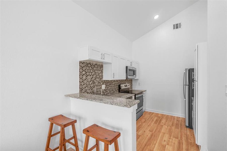 This is a view of the modern kitchen featuring white cabinetry, stainless steel appliances, a mosaic tile backsplash, and granite countertops. There's a convenient breakfast bar for casual dining. The space is well-lit with a high ceiling and recessed lighting.