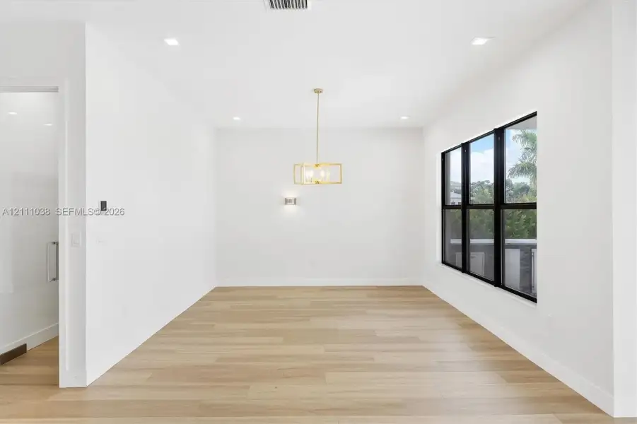 Open-concept interior featuring light wood-finish flooring, recessed lighting, a modern gold-tone geometric chandelier, and floor-to-ceiling windows with black framing