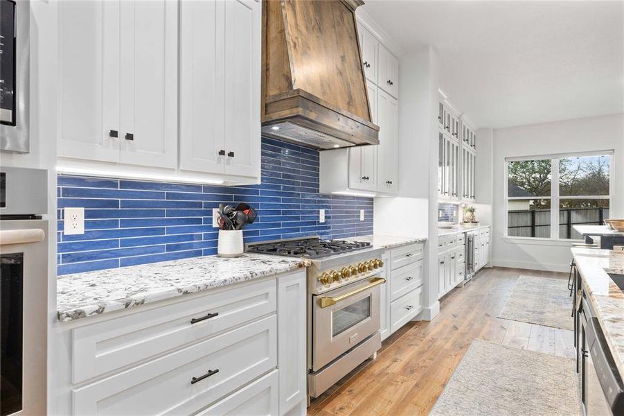 Kitchen featuring white cabinets, appliances with stainless steel finishes, light stone countertops, custom exhaust hood, and light wood-type flooring