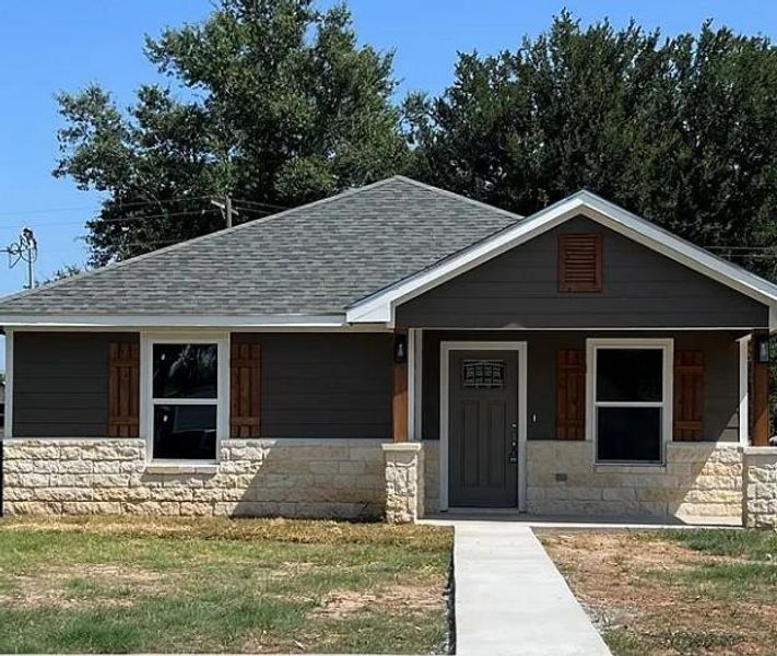 View of front of home with a porch, a shingled roof, stone siding, and a front lawn
