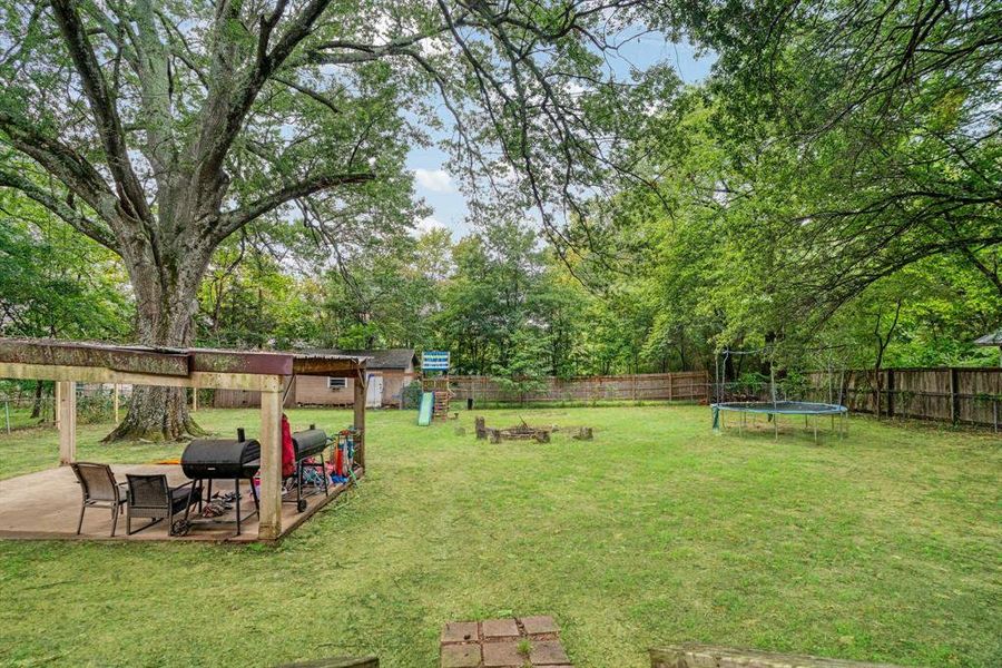 Fenced backyard with a trampoline, a playground, view of scattered trees, and a patio Fenced backyard with a trampoline, a playground, view of scattered trees, and a patio