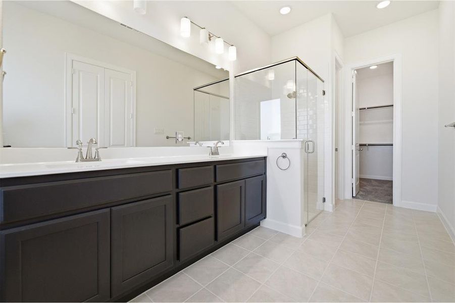 Bathroom featuring a dual vanity with dark wood-finish cabinetry, a large frameless mirror, a glass-enclosed shower with subway tile, and light-toned floor tiling