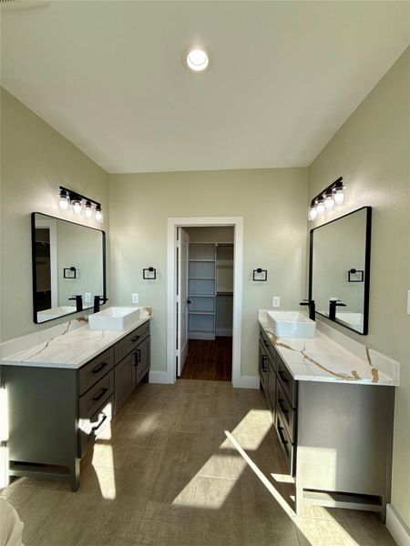 This photo shows a modern bathroom with dual vanities, each featuring quartz countertops and vessel sinks. The space includes sleek black fixtures, large mirrors, and symmetrical lighting. A walk-in closet is visible through an open door at the far end. This photo shows a modern bathroom with dual vanities, each featuring quartz countertops and vessel sinks. The space includes sleek black fixtures, large mirrors, and symmetrical lighting. A walk-in closet is visible through an open door at the far end.