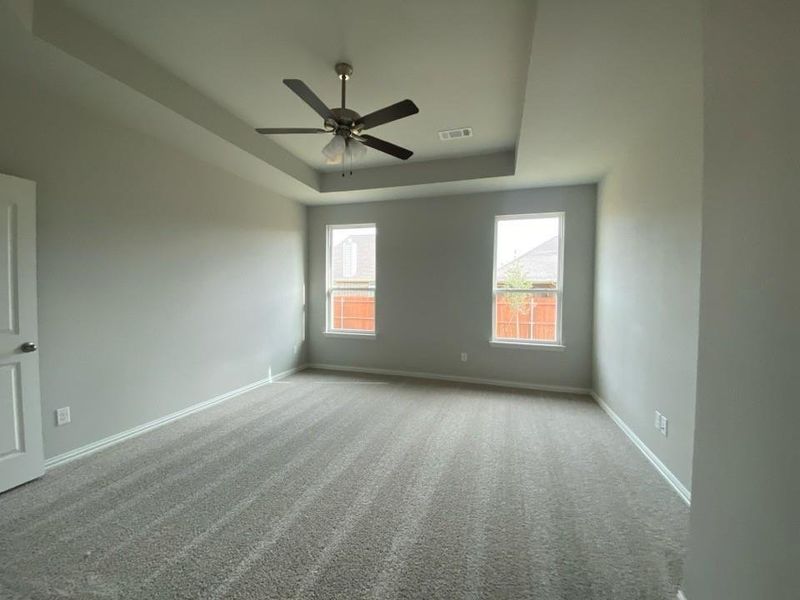 Spacious room featuring a tray ceiling with a ceiling fan, neutral wall paint, two windows, and gray carpeting Spacious room featuring a tray ceiling with a ceiling fan, neutral wall paint, two windows, and gray carpeting