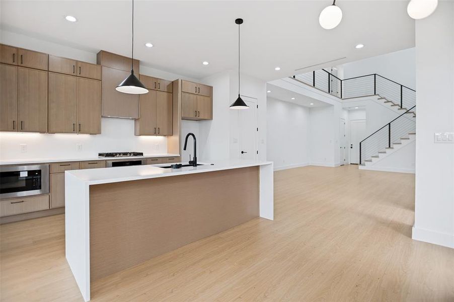 Modern kitchen featuring light wood-finish cabinetry, stainless steel appliances, a white countertop island with pendant lighting, and light wood-finish flooring
