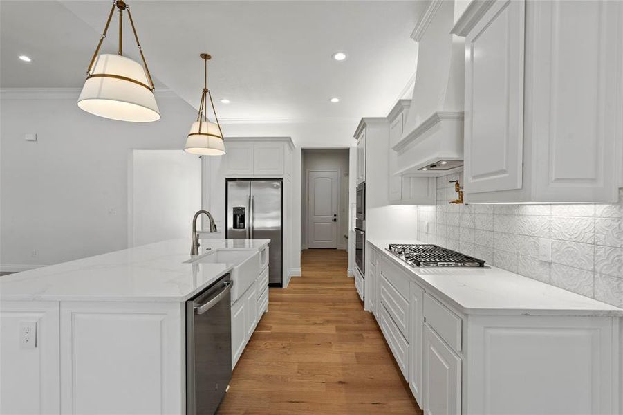 Kitchen with appliances with stainless steel finishes, light stone counters, custom range hood, light hardwood / wood-style flooring, and hanging light fixtures