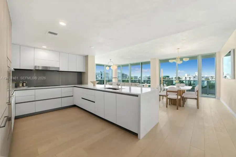 Contemporary kitchen featuring white handleless cabinetry, a substantial quartz-topped center island with integrated sink, stainless steel range hood, and light-toned wood-finish flooring, complemented by floor-to-ceiling glass wall panels Contemporary kitchen featuring white handleless cabinetry, a substantial quartz-topped center island with integrated sink, stainless steel range hood, and light-toned wood-finish flooring, complemented by floor-to-ceiling glass wall panels
