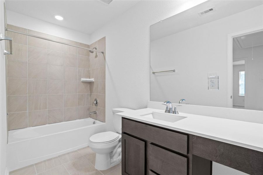 The secondary bathroom showcases tile flooring, a bath/shower combo with Daltile tile surround, a vanity with knee space and stunning light countertop, a mirror, sleek fixtures, and contemporary finishes throughout.