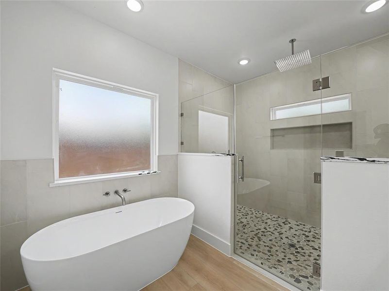 Freestanding soaking tub with wall-mounted faucet, alongside a glass-enclosed shower featuring a rainfall showerhead, pebble-tiled floor, and recessed shelf Freestanding soaking tub with wall-mounted faucet, alongside a glass-enclosed shower featuring a rainfall showerhead, pebble-tiled floor, and recessed shelf