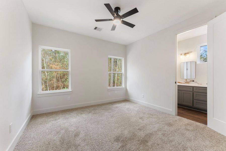 This photo shows a bright bedroom with soft carpeting, two windows offering natural light,  and an attached bathroom featuring modern fixtures and ample cabinet space.