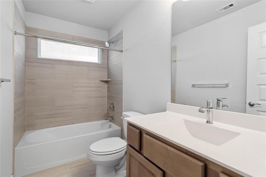 Secondary bath features tile flooring, bath/shower combo with tile surround, stained wood cabinets, beautiful light countertops, mirror, dark, sleek fixtures and modern finishes.