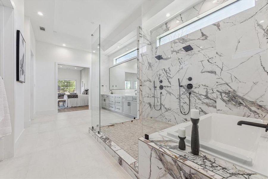 This primary bathroom features a luxurious design with exquisite surfaces, a frameless glass shower with dual shower heads, a large soaking tub, and a double vanity with ample storage.