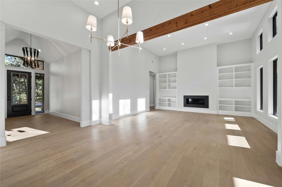 Unfurnished living room with light hardwood / wood-style flooring, a towering ceiling, beam ceiling, built in shelves, and an inviting chandelier