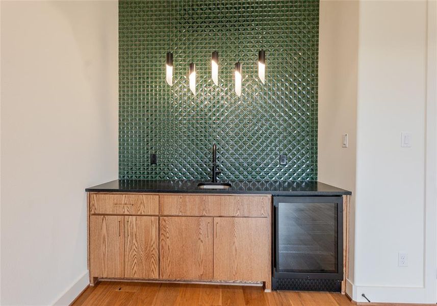 Indoor wet bar with wine cooler, light wood-style floors, dark stone countertops, hanging light fixtures, and light brown cabinets