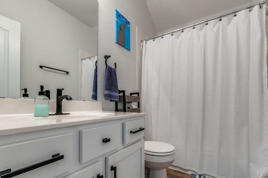 This modern bathroom features a clean design with white cabinetry, black fixtures, and a full shower with a white curtain. A simple wall decoration adds a touch of color, and the space is well-lit and inviting.