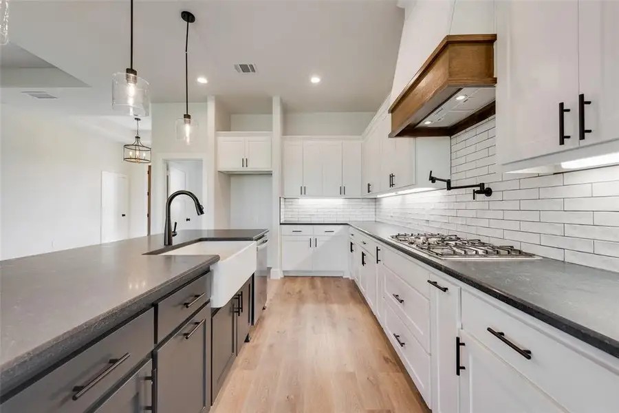 Kitchen featuring a large island with a farmhouse sink, light wood-finish flooring, white cabinetry with dark hardware, a white subway tile backsplash, and a gas cooktop with a wood-accented range hood