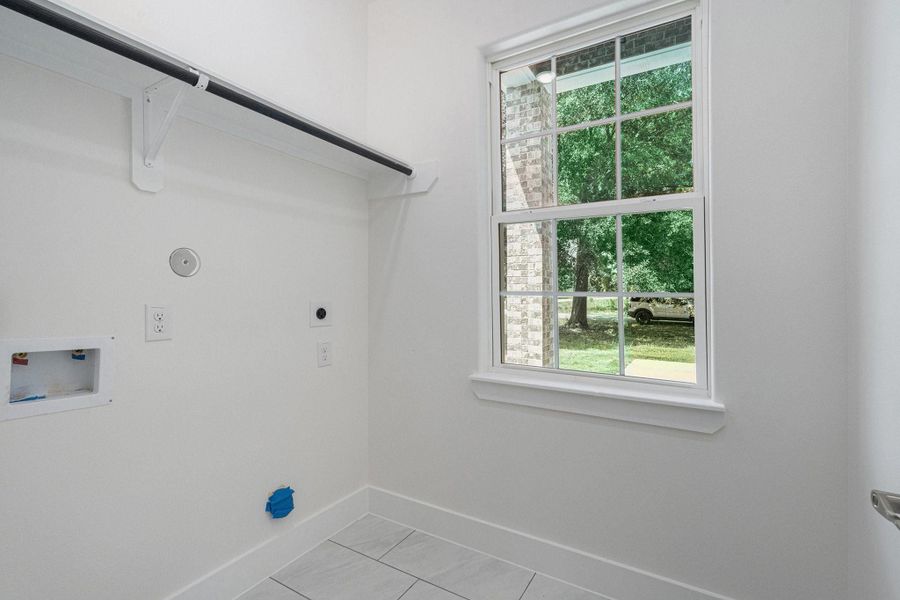 This photo shows a bright laundry room with white walls, a window providing natural light, utility hookups, and a shelf with a hanging rod. This photo shows a bright laundry room with white walls, a window providing natural light, utility hookups, and a shelf with a hanging rod.
