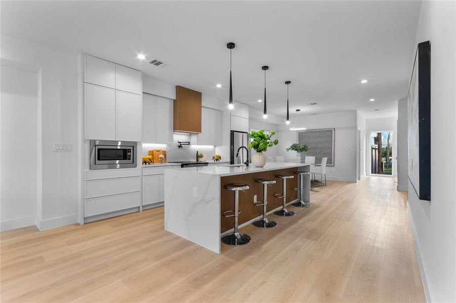 Gourmet kitchen featuring a waterfall island with integrated seating, stainless steel microwave, light wood-finish flooring, and pendant lighting Gourmet kitchen featuring a waterfall island with integrated seating, stainless steel microwave, light wood-finish flooring, and pendant lighting
