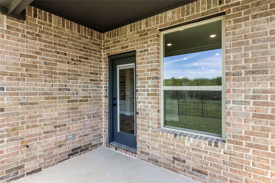 Covered back patio with brick exterior, dark blue door, and large window overlooking fenced yard Covered back patio with brick exterior, dark blue door, and large window overlooking fenced yard