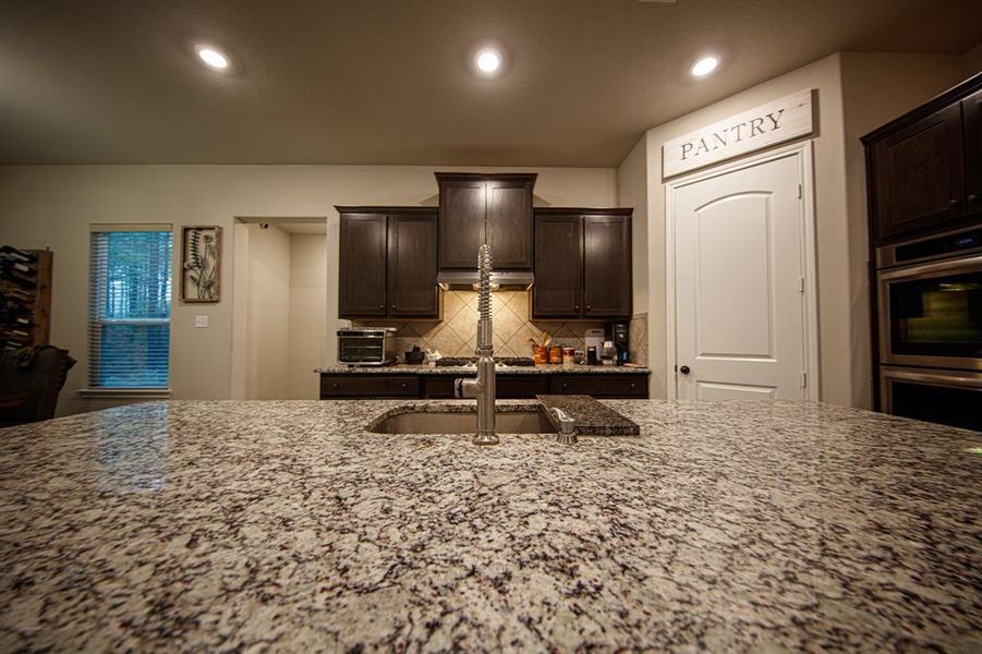 This kitchen features a spacious granite island with a sink, dark wood cabinetry, and stainless steel appliances. It includes a pantry, recessed lighting, and a cozy, modern atmosphere.