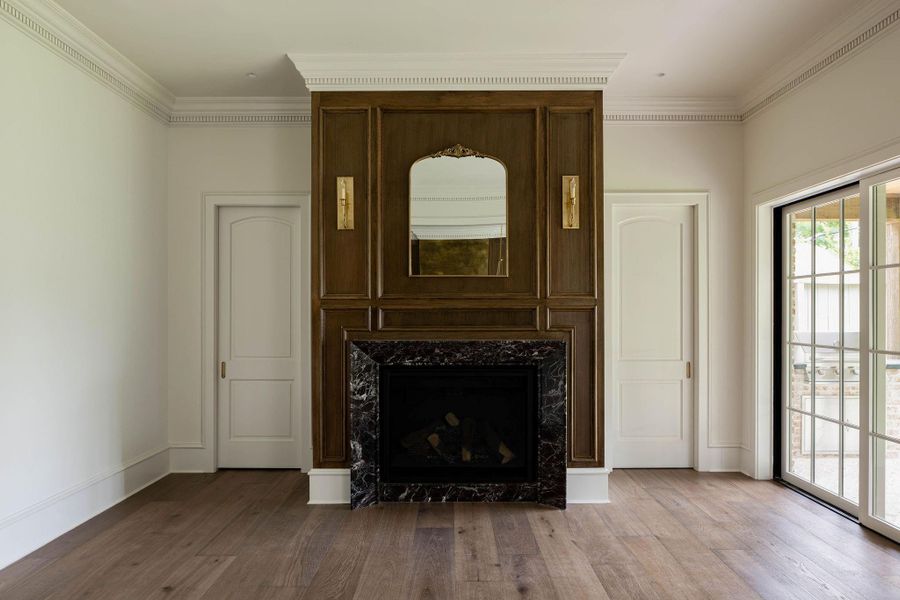 Living room anchored by a floor-to-ceiling solid wood paneled fireplace surround with custom raised paneling, a dark veined marble surround and hearth, brass wall sconces, and detailed dentil crown molding. Flanked by two arched-top passage doors and a multi-panel glass door opening to the back patio. Living room anchored by a floor-to-ceiling solid wood paneled fireplace surround with custom raised paneling, a dark veined marble surround and hearth, brass wall sconces, and detailed dentil crown molding. Flanked by two arched-top passage doors and a multi-panel glass door opening to the back patio.