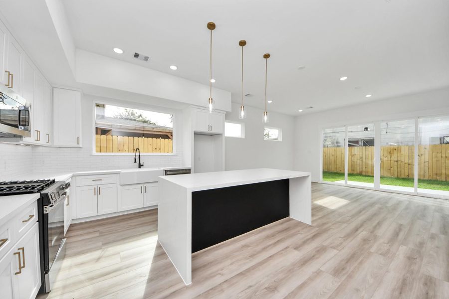 This photo showcases a modern, open-concept kitchen and living space. The kitchen features sleek white cabinetry, a central island with pendant lighting, and stainless steel appliances. Large windows and sliding doors provide ample natural light and lead to a fenced backyard. The light wood flooring enhances the bright, airy feel.