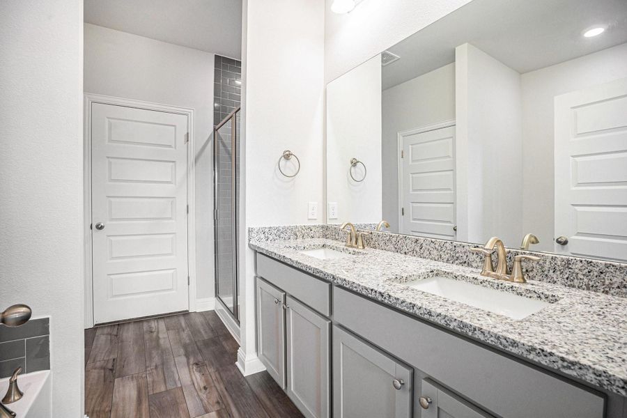 This primary bathroom features a spacious double vanity with granite countertops and modern fixtures. It has a sleek glass-enclosed shower, stylish gray cabinetry, and elegant luxury vinyl plank flooring, creating a fresh and contemporary feel.