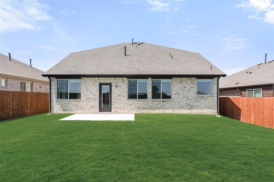 Rear exterior of single-story brick home with large windows, concrete patio, and fenced green backyard