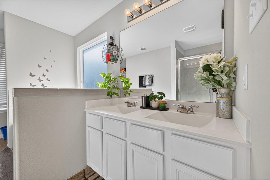This bathroom features a double sink vanity with white cabinets, a large mirror, modern lighting, and decorative touches like plants and wall accents. A frosted window provides natural light, and a walk-in shower is visible in the background.