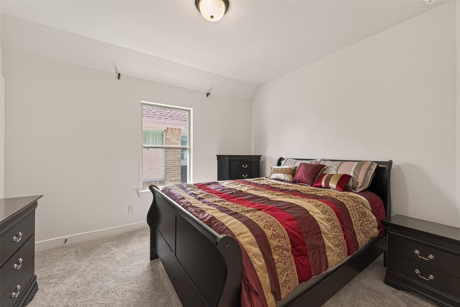 Cozy bedroom with plush carpet, featuring a large bed with colorful bedding. The room includes matching dark wood furniture and a window providing natural light. Simple and inviting space.