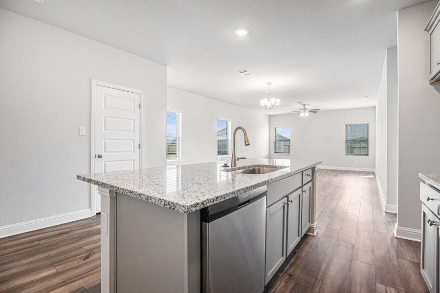 This photo showcases a modern kitchen with a spacious open layout. It features a large granite island with a sink, stainless steel appliances, and luxury vinyl plank flooring. The area is well-lit with ample natural light from multiple windows, leading into a living space with a modern light fixture and ceiling fan.