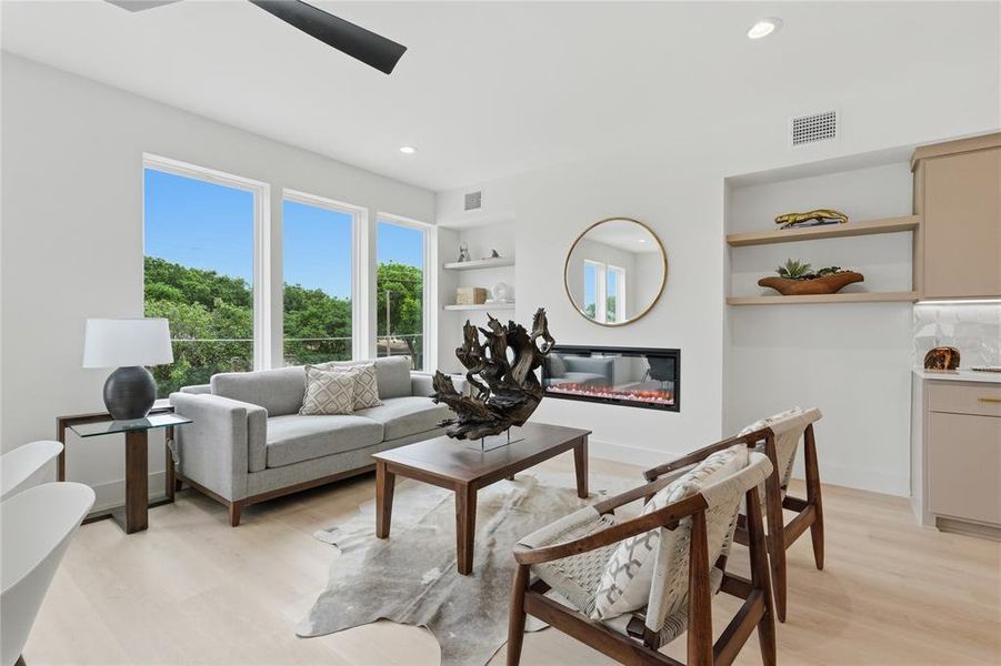 Light wood-finish flooring complements a modern living space featuring three large windows, a sleek linear electric fireplace, and recessed lighting Light wood-finish flooring complements a modern living space featuring three large windows, a sleek linear electric fireplace, and recessed lighting