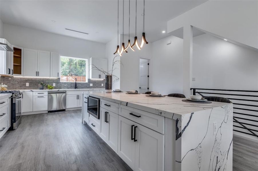 Kitchen with hanging light fixtures, wood-type flooring, appliances with stainless steel finishes, light stone countertops, and a center island