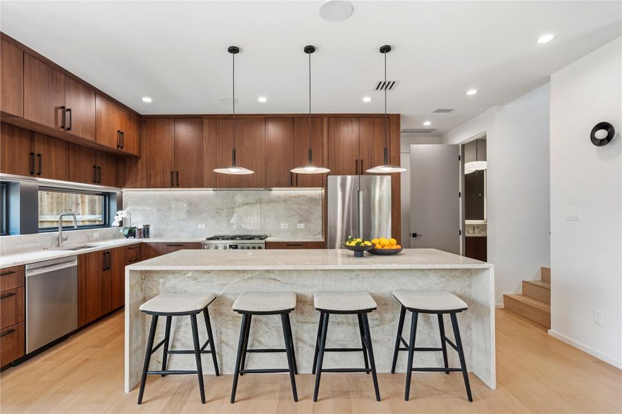 A pristine kitchen features sleek wood cabinetry, marble counters, stainless appliances, and pendant lighting illuminating a generous island.