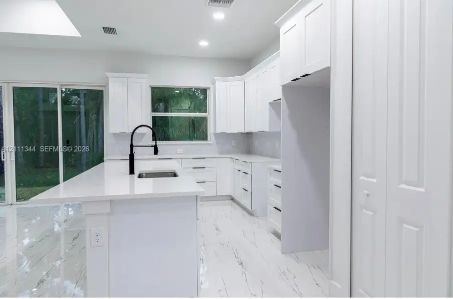 Kitchen featuring white shaker-style cabinetry, a quartz-topped island with an undermount stainless steel sink, matte black hardware, stone-patterned tile flooring, and a glass-paneled sliding door Kitchen featuring white shaker-style cabinetry, a quartz-topped island with an undermount stainless steel sink, matte black hardware, stone-patterned tile flooring, and a glass-paneled sliding door